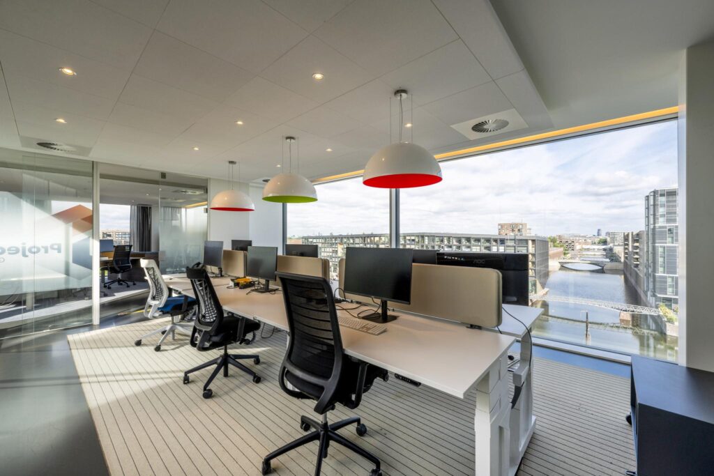 Modern office space on Krijn Taconiskade with workstations, colorful pendant lights, and a canal view through large windows.